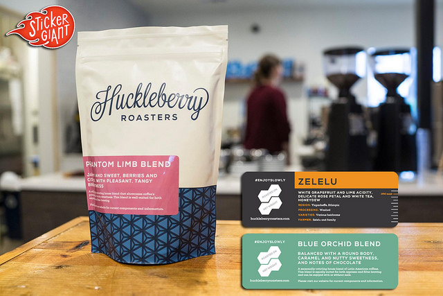 Custom-coffee-labels-printed-by-StickerGiant-for-Huckleberry-Roasters-with-a-bag-of-coffee-and-a-coffee-shop-in-the-background