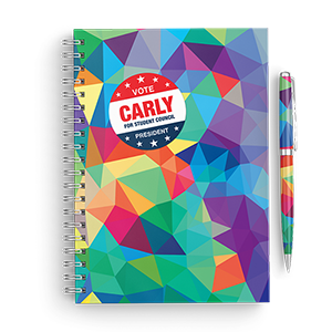 A mosaic colored spiral notebook with a Vote Carly sticker printed by StickerGiant applied to the front and a mosaic colored pen laying next to the notebook